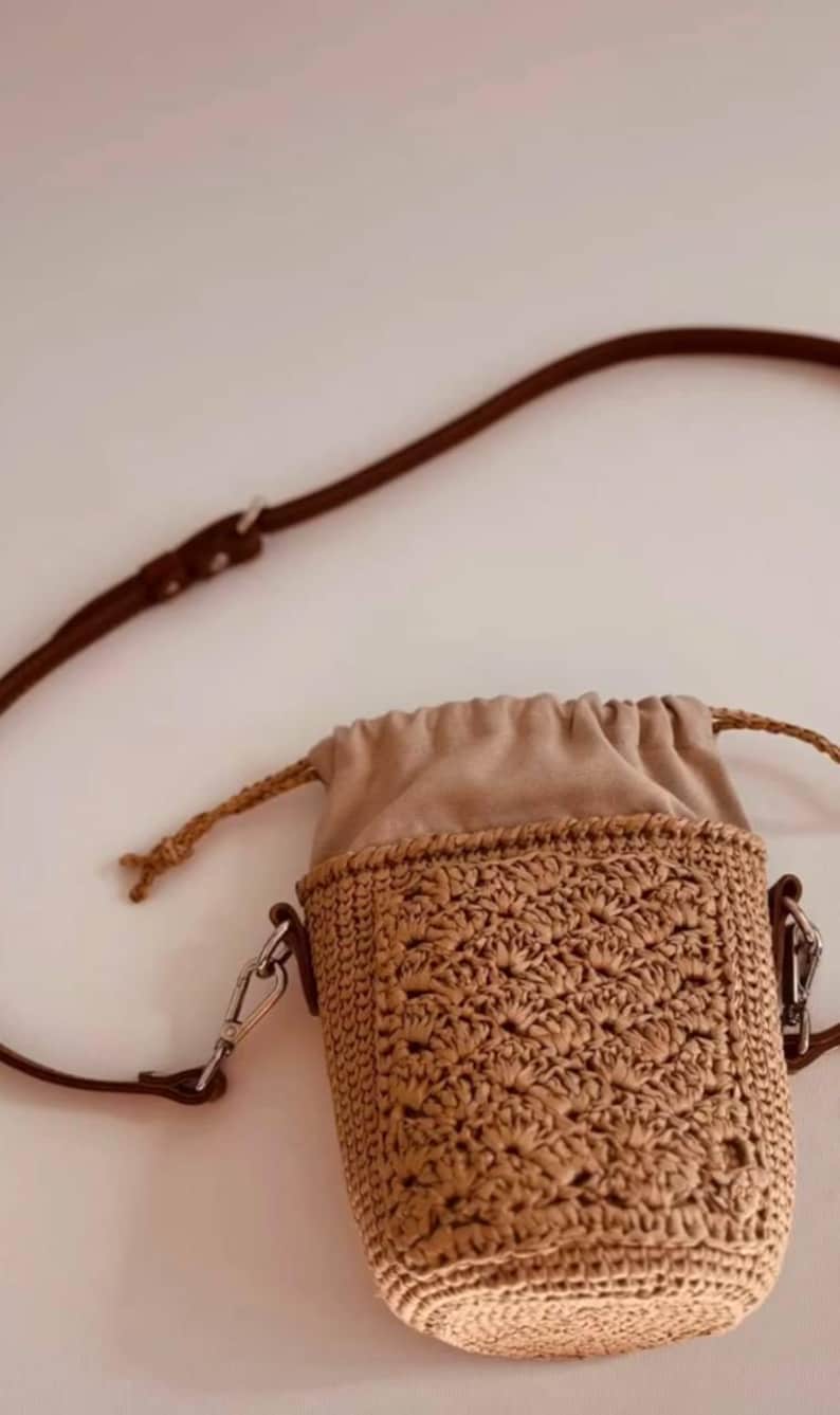 May include: A small cylindrical crossbody bag featuring a tan crocheted raffia body with a textured shell-like pattern. The bag has a beige fabric drawstring closure at the top, a thin brown leather strap with silver metal hardware, and side clips.