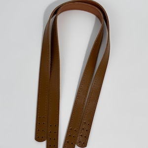 May include: A pair of tan brown bag handles featuring a smooth finish and stitched edges. Each strap has four pre-punched holes at both ends for attachment, providing a clean and functional design for a handbag or tote replacement project.