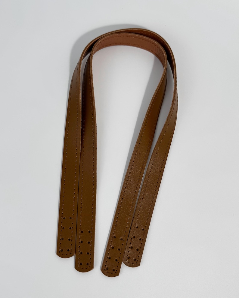 May include: A pair of tan brown bag handles featuring a smooth finish and stitched edges. Each strap has four pre-punched holes at both ends for attachment, providing a clean and functional design for a handbag or tote replacement project.