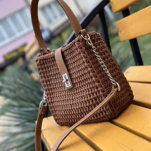 Puede incluir: Un bolso de mano de ganchillo marrón con forma rectangular estructurada, un asa superior y una correa de hombro desmontable con eslabones de cadena plateados. El bolso incluye una tira central vertical con un cierre giratorio de metal plateado.