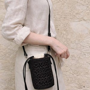 May include: A small, cylindrical black crochet crossbody bag with a drawstring closure. The bag features a textured, woven pattern and is attached to a thin, adjustable black strap. The top opening is secured with a light beige fabric lining.