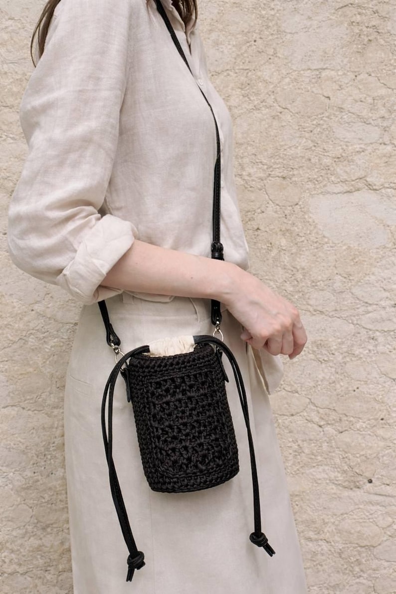 May include: A small, cylindrical black crochet crossbody bag with a drawstring closure. The bag features a textured, woven pattern and is attached to a thin, adjustable black strap. The top opening is secured with a light beige fabric lining.
