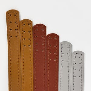 May include: A collection of leather straps in varying shades of brown and gray, arranged vertically against a white background. Each strap has a rounded end with stitched detailing and evenly spaced holes for adjustment. The straps are likely for a bag or other accessory.