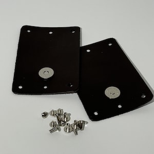 Op de afbeelding: Twee rechthoekige zwarte leren stukken, elk voorzien van een gecentreerde zilverkleurige magnetische drukknoop en vier voorgeponste gaten langs de randen. Een kleine hoop zilverkleurige metalen klinknagels en bevestigingsmiddelen ligt onder de stukken.