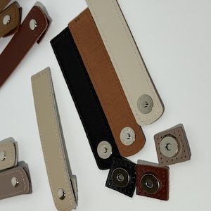 May include: A collection of rectangular and square leather-like straps and tabs in black, tan, beige, and brown colors. Each piece features a metallic silver-toned snap button fastener. The items are arranged in various orientations on a plain surface.