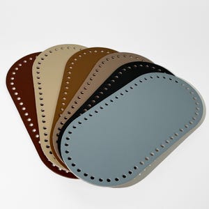 Könnte beinhalten: Eine Auswahl an ovalen Taschenböden in verschiedenen Farben, darunter Braun, Beige, Hellbraun, Schwarz und Blau. Jeder Boden hat gleichmäßig verteilte Löcher am Rand zum Annähen. Die Böden bestehen aus einem glatten, einfarbigen Material.