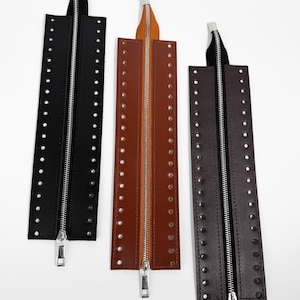 May include: Three leather zipper pulls in black, brown, and dark brown. Each pull features a metal zipper and is adorned with evenly spaced silver rivets along the edges. The pulls have a fabric loop at the top for attachment.