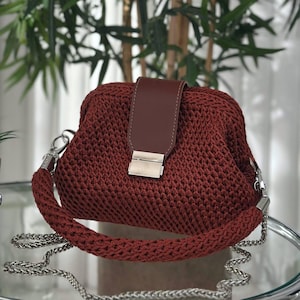May include: A burgundy crochet handbag with a brown leather flap and silver-tone hardware. The bag features a braided handle and a silver chain strap. The bag is approximately 10 inches wide.