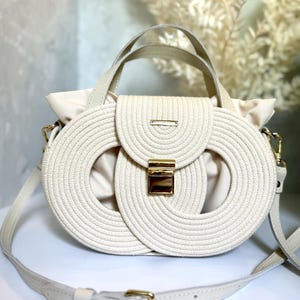 May include: A cream-colored handbag featuring a structured exterior made of coiled rope in an oval pattern. The bag includes a central gold-tone metal clasp, a top handle, and an adjustable shoulder strap with gold-tone hardware accents.