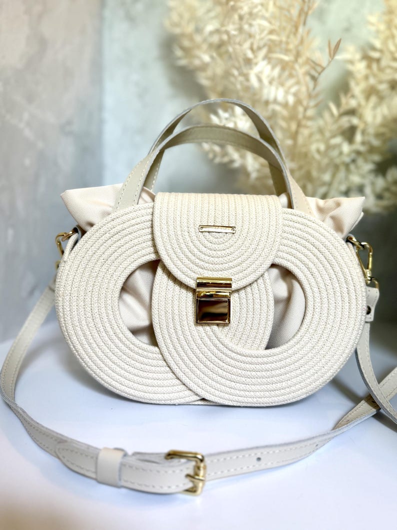 May include: A cream-colored handbag featuring a structured exterior made of coiled rope in an oval pattern. The bag includes a central gold-tone metal clasp, a top handle, and an adjustable shoulder strap with gold-tone hardware accents.