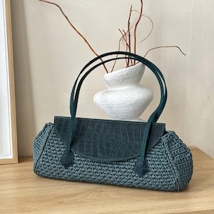 May include: A teal handbag featuring a textured, crocheted body and a smooth, dark teal flap with a crocodile-skin pattern. The bag includes two matching teal shoulder straps attached to the front flap with small, pointed, textured leather tabs.