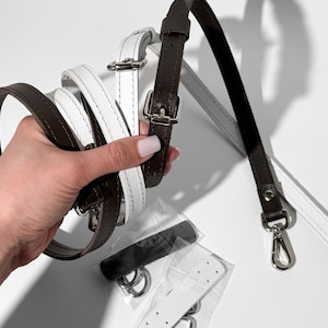 K&ouml;nnte beinhalten: Zwei verstellbare Taschenriemen aus dunkelbraunem und wei&szlig;em Leder, jeweils mit silberfarbenen Metallschnallen und Karabinerhaken. Zwei kleine, separate Lederlaschen mit silbernen D-Ringen sind in einer transparenten Kunststoffverpackung enthalten.