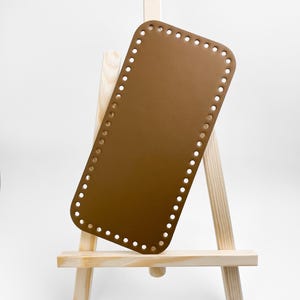 Puede incluir: Base de bolso rectangular marrón con agujeros espaciados alrededor del perímetro, exhibida en un pequeño caballete de madera. La base es de color marrón mate sólido. El caballete es de madera clara.