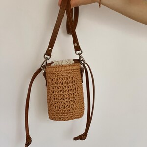 May include: A cylindrical woven straw bucket bag in a light tan color featuring a textured crochet pattern, a cream-colored fabric drawstring closure at the top, and a thin brown leather shoulder strap attached with silver-toned metal hardware.