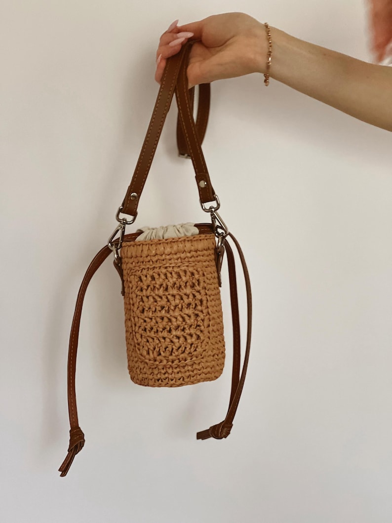 May include: A cylindrical woven straw bucket bag in a light tan color featuring a textured crochet pattern, a cream-colored fabric drawstring closure at the top, and a thin brown leather shoulder strap attached with silver-toned metal hardware.