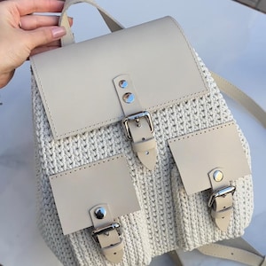 May include: A cream-colored backpack featuring a crocheted body and smooth, light-colored leather-like accents. The bag includes a main flap with a silver-toned metal buckle closure and two smaller front pockets, each secured with a matching buckle.
