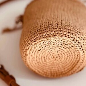 May include: A cylindrical woven straw bag in a light tan color featuring a circular base with a concentric pattern. The bag includes a thin brown leather strap with visible stitching and a metal buckle, attached to the side of the woven body.