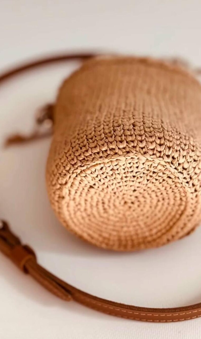 May include: A cylindrical woven straw bag in a light tan color featuring a circular base with a concentric pattern. The bag includes a thin brown leather strap with visible stitching and a metal buckle, attached to the side of the woven body.