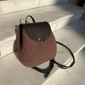 Puede incluir: Una mochila de ganchillo marrón con una solapa lisa de color marrón oscuro y un cierre de metal plateado. El cuerpo del bolso presenta un patrón de punto denso y texturizado, e incluye correas finas y ajustables de color marrón oscuro.