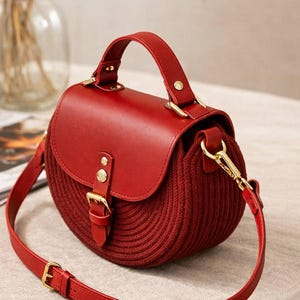 May include: A red handbag with a rounded shape, featuring a knitted body and a leather flap. It has a top handle and a long, adjustable shoulder strap. Gold-coloured hardware accents the bag, including the clasp and buckle.