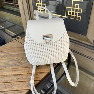 May include: A white backpack featuring a textured, woven body and a smooth, solid white flap closure. The bag includes a silver-toned metal clasp on the front, a top handle, and adjustable shoulder straps, all in a matching white color.