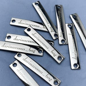 May include: A collection of shiny, silver rectangular tags with rounded ends. Each tag has a small hole at each end and the engraved text "darinavesna_bag" in a cursive font. The tags are scattered on a light blue surface.