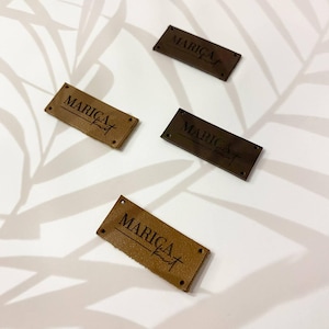 May include: Four rectangular labels in varying shades of brown, ranging from light tan to dark chocolate. Each label features the text MARICA knit embossed in a dark font and includes small pre-punched holes at the corners for attachment purposes.
