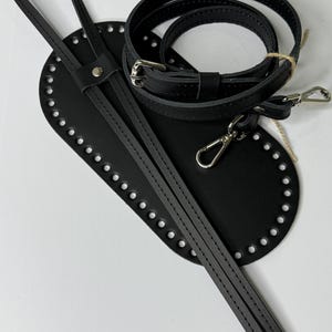 May include: A black oval-shaped leather base with pre-punched holes along the perimeter, accompanied by a long black leather strap with a silver metal buckle and two silver swivel lobster claw clasps for attachment to a bag or accessory.