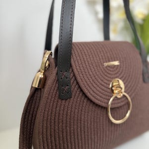 May include: A brown handbag constructed from coiled cord material featuring dark brown leather straps, a gold-toned circular ring clasp on the front flap, and gold metal hardware accents. The bag has a structured, rounded shape with visible stitching.