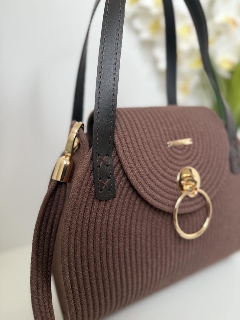 May include: A brown handbag constructed from coiled cord material featuring dark brown leather straps, a gold-toned circular ring clasp on the front flap, and gold metal hardware accents. The bag has a structured, rounded shape with visible stitching.
