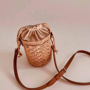 May include: A cylindrical bucket bag featuring a tan crocheted exterior with a textured floral pattern. The bag has a beige satin drawstring closure at the top and a thin, adjustable brown leather shoulder strap with a silver metal buckle.
