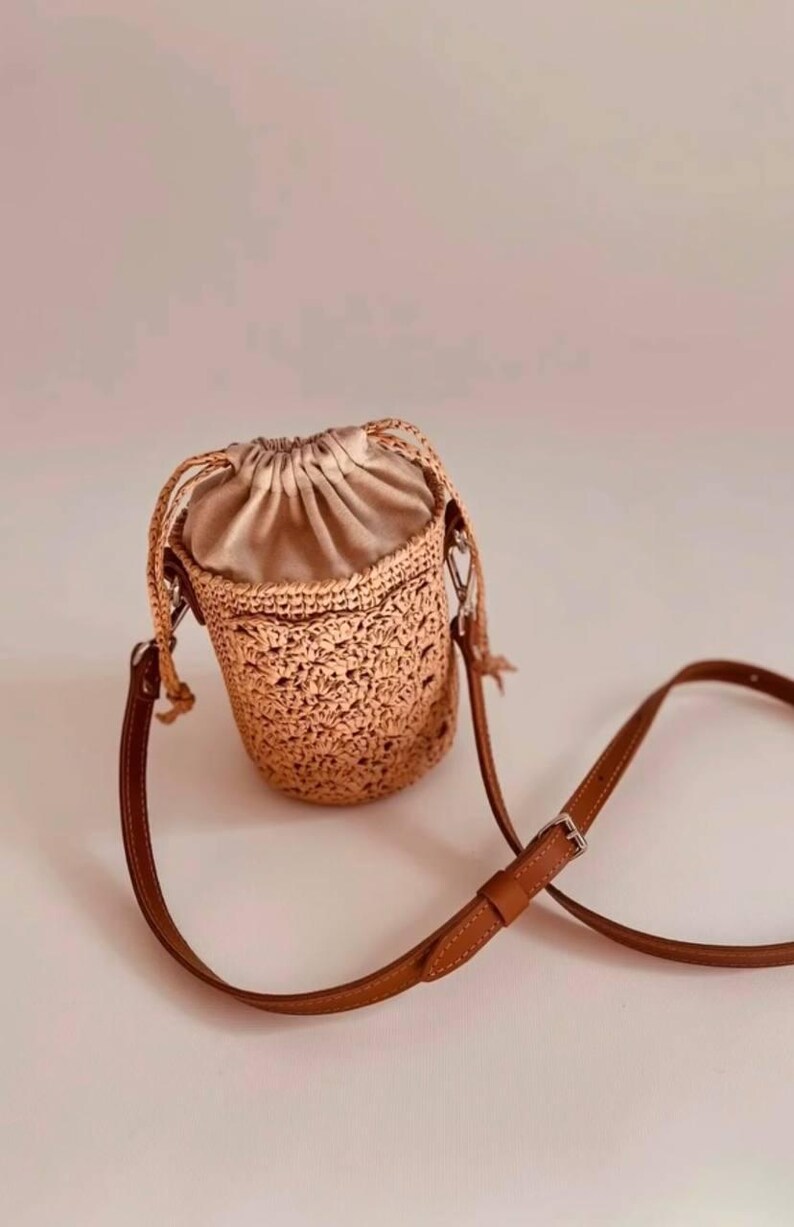 May include: A cylindrical bucket bag featuring a tan crocheted exterior with a textured floral pattern. The bag has a beige satin drawstring closure at the top and a thin, adjustable brown leather shoulder strap with a silver metal buckle.