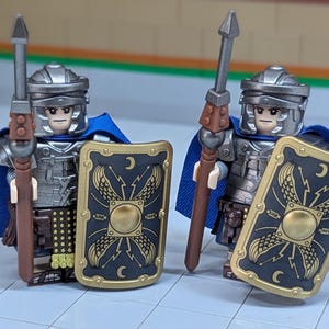 May include: Two toy Roman legionaries. Each figure wears a silver helmet, armor, and a blue cape. They hold a spear and a black shield with gold accents and a crescent moon design. The figures are standing on a light gray surface.