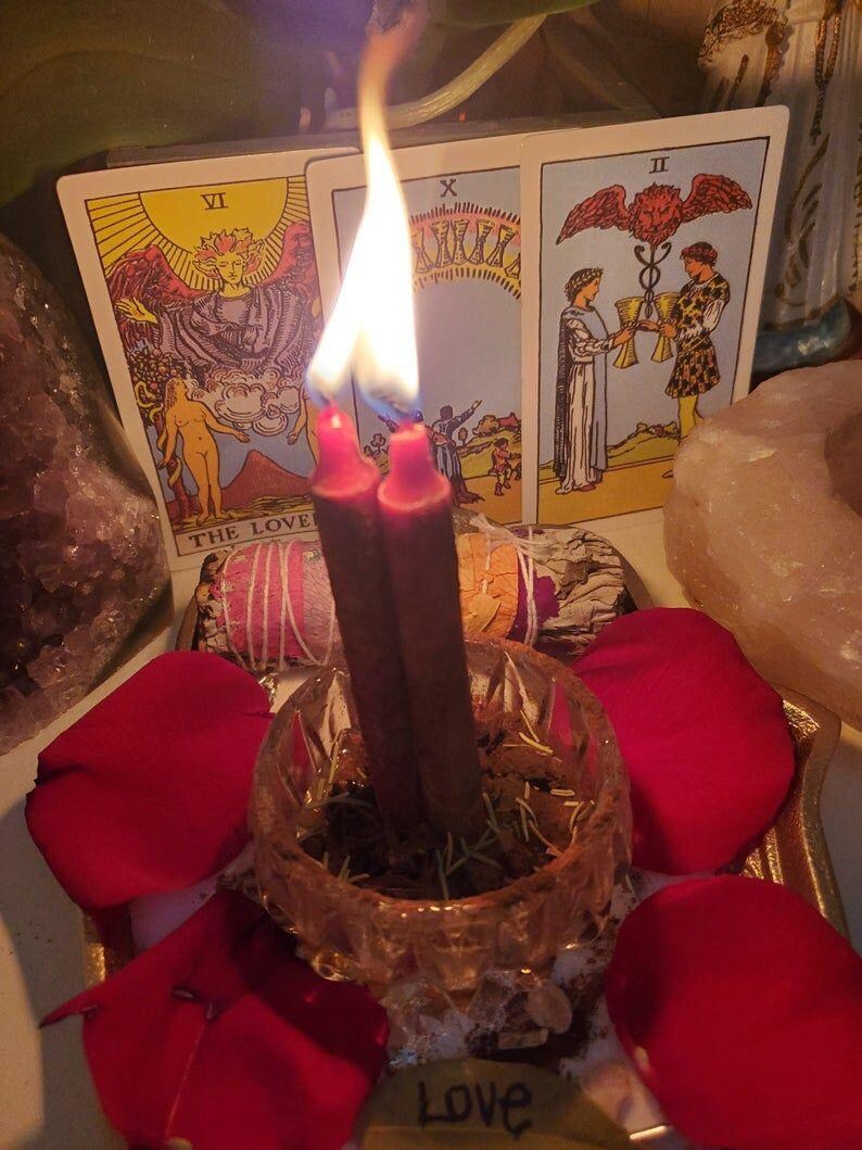 May include: A still-life image featuring lit red candles in a glass bowl, surrounded by red rose petals. Tarot cards with the words "THE LOVE" and "Love" are visible. The scene evokes a spiritual or romantic theme.