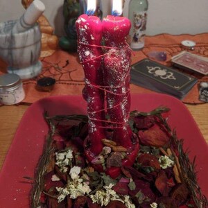 May include: Two red, lit candles tied together with red string, sitting on a red plate surrounded by dried herbs and flowers. The candles are in the shape of two figures. A mortar and pestle are in the background.
