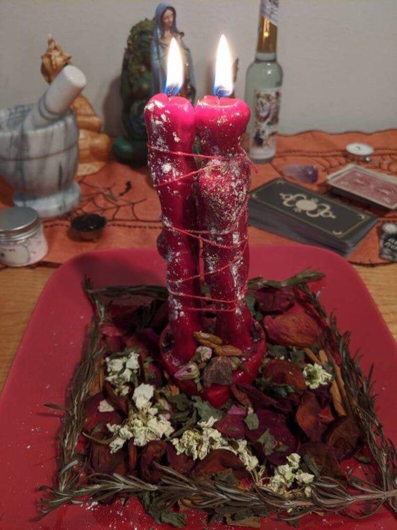 May include: Two red, lit candles tied together with red string, sitting on a red plate surrounded by dried herbs and flowers. The candles are in the shape of two figures. A mortar and pestle are in the background.
