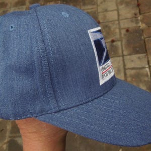 Könnte beinhalten: Blaue Baseballkappe aus Denim mit dem Logo der United States Postal Service auf der Vorderseite. Die Kappe hat einen gebogenen Schirm und eine strukturierte Krone. Das Logo zeigt einen weißen Adler und die Worte "UNITED POSTAL SERVICE".