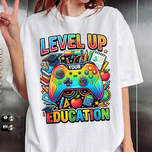 May include: A white t-shirt featuring a colorful graphic with the text "LEVEL UP YOUR EDUCATION". The design includes a rainbow-colored video game controller, graduation caps, books, pencils, apples, a calculator, and a globe in a vibrant style.