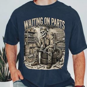 May include: Navy blue t-shirt featuring a detailed graphic of a skeleton mechanic sitting on tires in a garage, holding a wrench. The text "WAITING ON PARTS GARAGE" is above the skeleton. Tools are scattered around the tires.