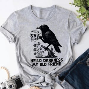 May include: A light gray t-shirt featuring a black illustration of a raven perched on a tree branch, holding a skull-shaped mug. The text "HELLO DARKNESS MY OLD FRIEND" is printed below the raven. The shirt is knotted at the waist.
