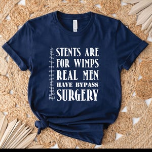 May include: Navy blue t-shirt with white text that reads "STENTS ARE FOR WIMPS REAL MEN HAVE BYPASS SURGERY". The shirt is made of a soft material and has short sleeves. The shirt is knotted at the bottom.
