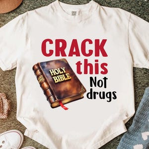 May include: White t-shirt with the text "CRACK this Not drugs" in red and black. The shirt features a graphic of a brown Holy Bible with gold lettering and a red ribbon bookmark. The shirt is made of a soft, comfortable material.