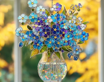 May include: A stained glass suncatcher featuring a bouquet of blue and purple flowers in a clear glass vase. The flowers have yellow centers and green leaves, all outlined in dark metal. The vase contains clear water with iridescent light reflections.