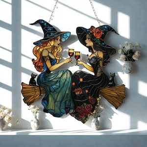 May include: Stained glass artwork featuring two witches toasting with wine glasses. They are seated on brooms, with black cats nearby. The witches wear pointed hats and dresses in shades of blue, green, and black, with floral accents. The artwork is suspended by chains.
