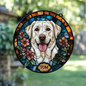 May include: Round stained glass window featuring a smiling yellow Labrador Retriever with its tongue out. The dog is surrounded by blue and orange glass, with pink flowers and green leaves. The name "TOM" is displayed on a small plaque.