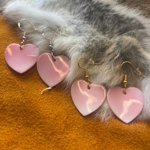 May include: Two pairs of heart-shaped earrings. One pair features light pink hearts with silver-toned metal hooks, while the other pair features pink-to-white gradient hearts with gold-toned metal hooks. Each earring has a smooth, glossy finish.
