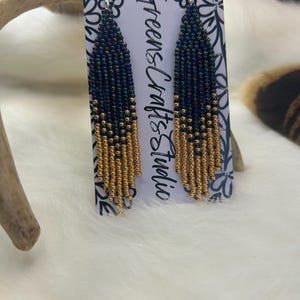 May include: Handmade beaded earrings with a fringe design. The earrings feature a gradient of dark blue and gold beads, with a silver hook. The text "Greens & Crafts Studio" is printed on the backing card.