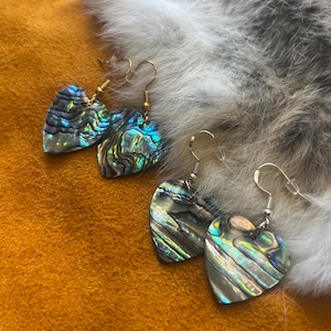 Pode incluir: Dois pares de brincos em forma de coração feitos de concha de abalone iridescente. Um par possui ganchos de metal dourado, enquanto o outro par possui ganchos de metal prateado. Cada brinco exibe um padrão natural e ondulado de tons azuis, verdes e roxos.