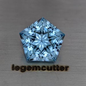 Puede incluir: Una gema facetada de color azul claro en forma de estrella. La gema tiene un patrón interno complejo y está colocada sobre una superficie de metal cepillado. El texto "legemcutter" es visible debajo de la gema.