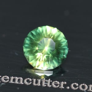 May include: A faceted, round gemstone with a vibrant green hue. The gem has a complex, multi-faceted design that reflects light, creating a sparkling effect. The gem is set against a dark background, highlighting its color and brilliance.
